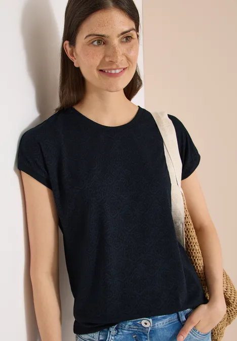 Shirt met burn-out look universal blue Persoon leunt tegen muur, draagt een zwart t-shirt en een strooien tas.