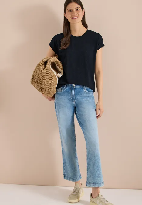 Shirt met burn-out look universal blue Vrouw voor beige muur met zwart shirt, lichtblauwe jeans, sneakers en gevlochten strooien tas.