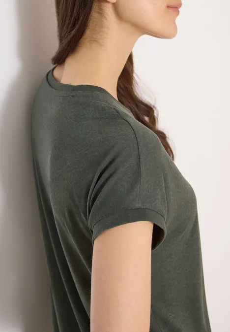 Jonge vrouw in zijaanzicht, leunt tegen een witte muur, draagt een donkergroen kort T-shirt, kijkt voor zich