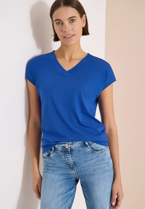 Frau in blauem V-Shirt und Jeans lehnt vor hellem Hintergrund.