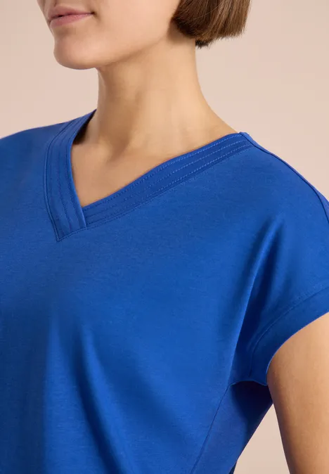 Nahaufnahme einer Person in royalblauem V-Ausschnitt-Shirt vor neutralem Hintergrund.