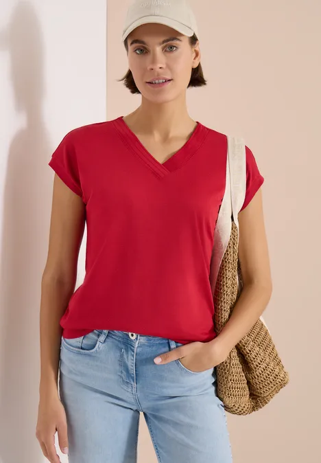 Vrouw draagt rood V-shirt, lichtblauwe jeans en rotan tas voor neutrale achtergrond.