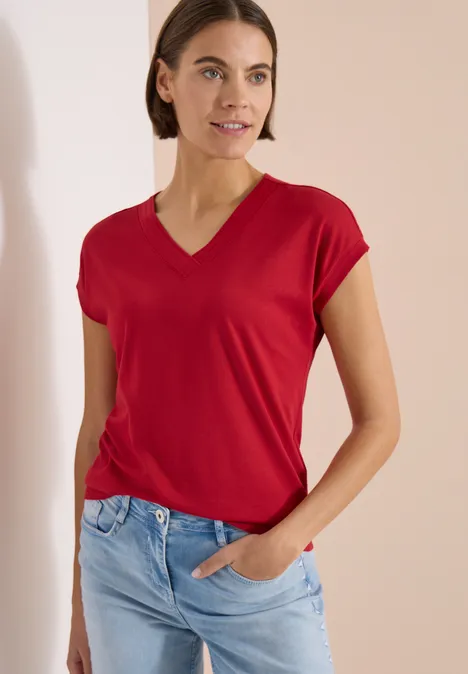 Vrouw met rood V-T-shirt en blauwe jeans staat met één hand in de zak voor een neutrale achtergrond.