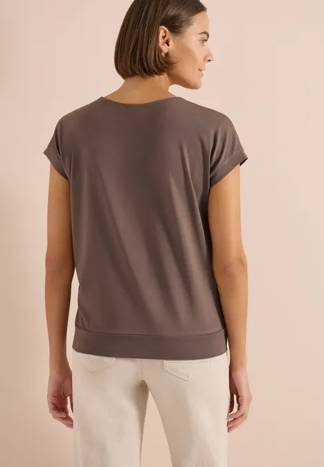 Shirt met korte mouwen en V-hals in effen kleur urban taupe Vrouw van achteren in losse taupekleurige top met korte mouwen en lichte broek, kort bruin haar.
