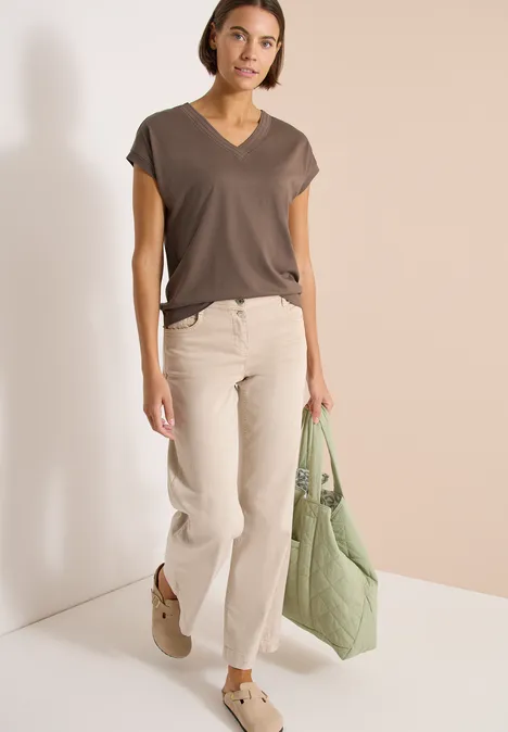 Shirt met korte mouwen en V-hals in effen kleur urban taupe Vrouw in donkergrijs V-hals shirt, lichte wijde broek, bruine loafers en groene gewatteerde tas voor een beige wand.