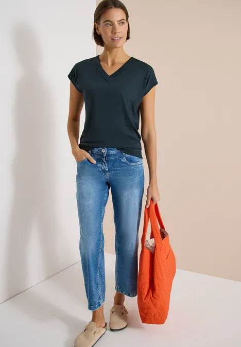 Shirt met korte mouwen en V-hals in effen kleur deep petrol blue Persoon voor een lichte muur, met een zwart T-shirt, blauwe spijkerbroek, beige schoenen en een oranje tas.