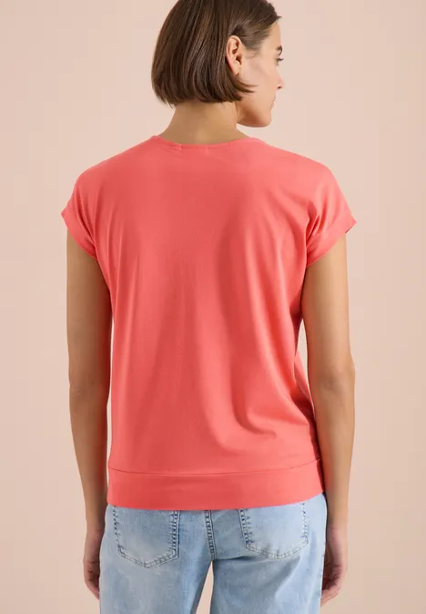 Rugaanzicht van een persoon in een koraalkleurig T-shirt en lichtblauwe jeans voor een neutrale achtergrond.