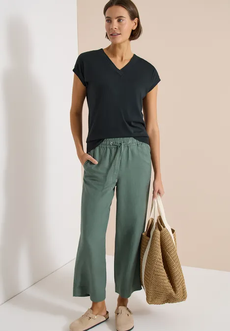 Een vrouw draagt een zwart t-shirt, wijde groene broek en houdt een grote gevlochten tas vast.