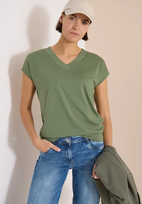 Person trägt olivgrünes V-Neck-Shirt, blaue Jeans, hält grüne Jacke lässig in der Hand.