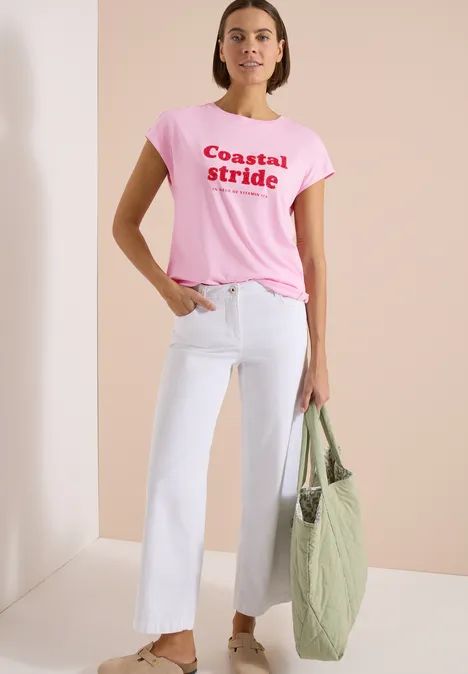 Shirt met boothals en tekstopdruk light blush rose Vrouw draagt roze T-shirt met "Coastal Stride", witte wijde cropped jeans en groene gewatteerde shopper.