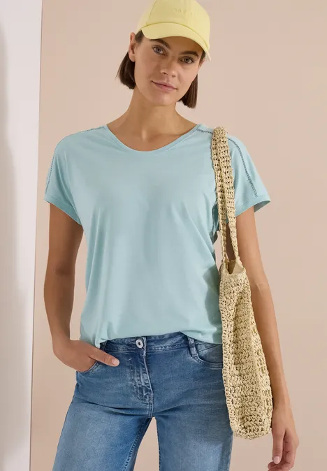 Shirt met V-hals en kanten detail crystal mint Een vrouw draagt een lichtblauw shirt, spijkerbroek en een strooien tas.