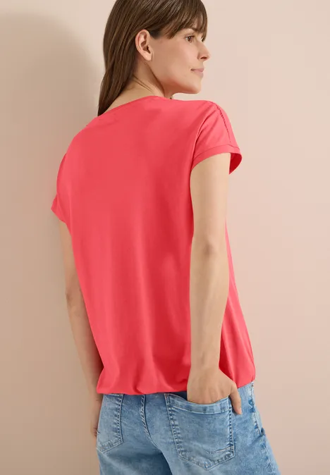 T-Shirt mit V-Neck und Spitzendetail coral Rückansicht einer Frau in rotem T-Shirt und Jeans, eine Hand in der Hosentasche.