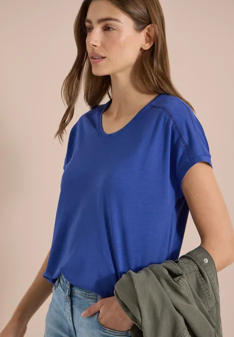 Frau mit verschwommenem Gesicht trägt blaues T-Shirt, hält grüne Jacke in der Hand.