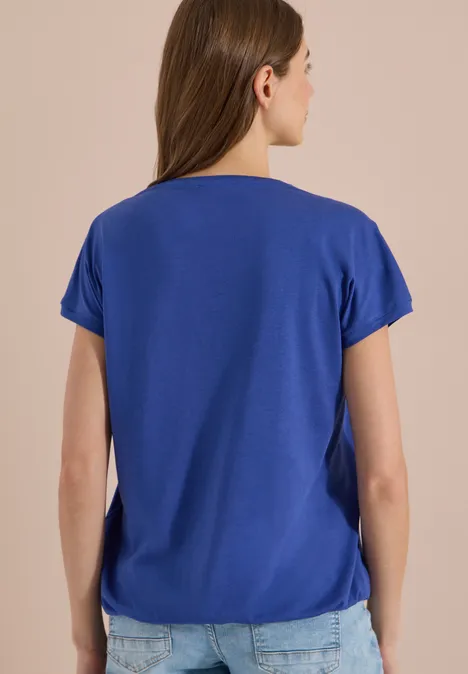 Rückansicht einer Frau mit schulterlangem Haar, die ein blaues kurzärmeliges T-Shirt und Jeans trägt.