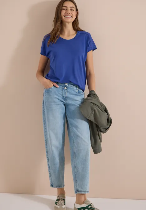 Frau in blauem T-Shirt und hellblauer Jeans, hält eine grüne Jacke vor beigem Hintergrund.