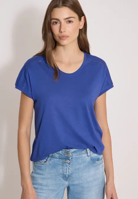 Frau trägt blaues T-Shirt und Jeans, steht vor neutralem Hintergrund.