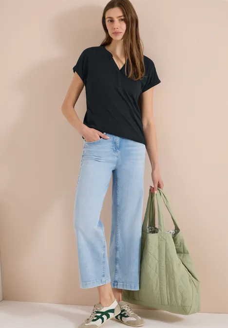 Frau in schwarzem T-Shirt, hellblauen Jeans, weißen Sneakern mit grünem Detail, trägt große grüne Tasche.