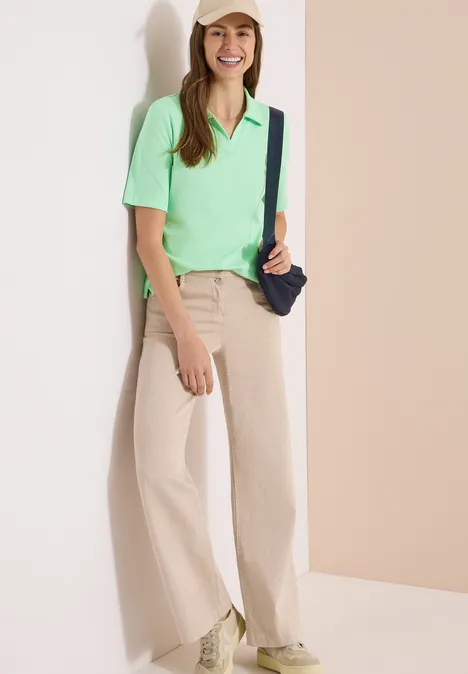 Vrouw leunt tegen lichte muur met mintgroene polo, beige wijdvallende broek, witte sneakers en zwarte tas.