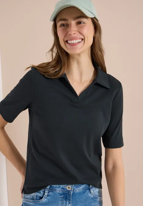 Vrouw met zwart T-shirt, blauwe jeans en lichtgekleurde pet tegen een beige achtergrond.