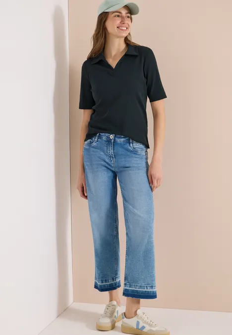 Vrouw in zwart poloshirt, lichtblauwe cropped jeans, pet en sneakers voor een beige achtergrond.
