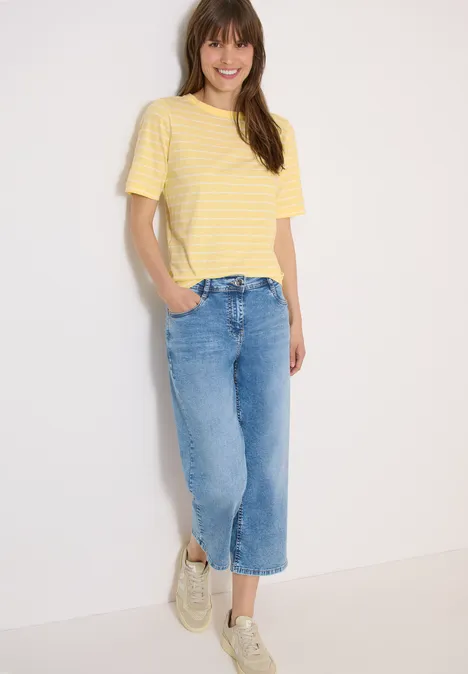Vrouw in geel-wit gestreept T-shirt en wijd, enkellange jeans met zijsplit, witte sneakers.