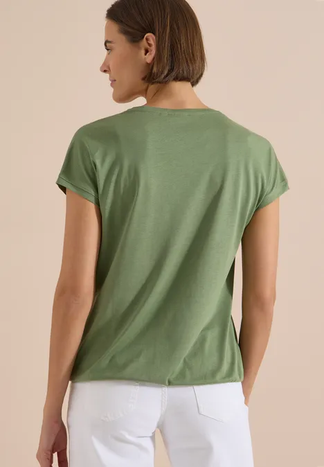 Eine Frau von hinten in grünem T-Shirt und weißer Hose vor schlichtem Hintergrund.