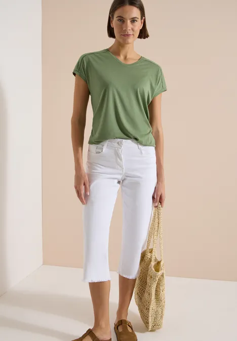 Frau trägt grünes T-Shirt, weiße Capri-Hosen und hält eine geflochtene Strohtragetasche.