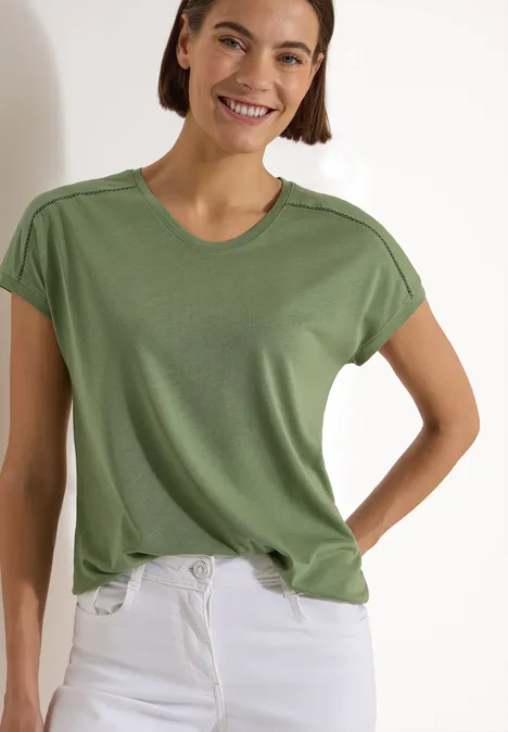 Eine Person trägt ein schlichtes grünes T-Shirt und weiße Jeans, Oberkörper sichtbar.