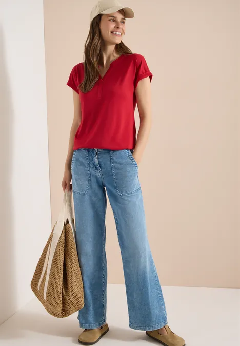 Kurzarm Shirt im Tunika-Look tamarillo red Frau in rotem T-Shirt, weiten blauen Jeans und geflochtener Strohtasche vor neutralem Hintergrund.