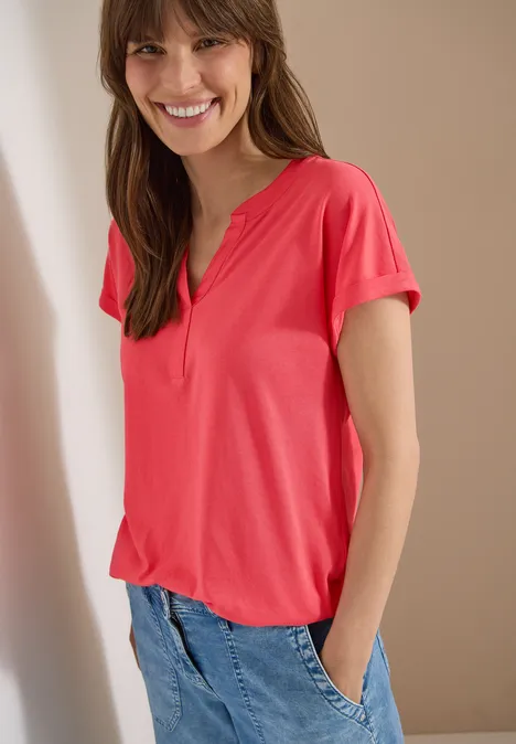 Kurzarm Shirt im Tunika-Look coral Frau im pinken T-Shirt mit Henley-Kragen und Jeans vor hellem Hintergrund.