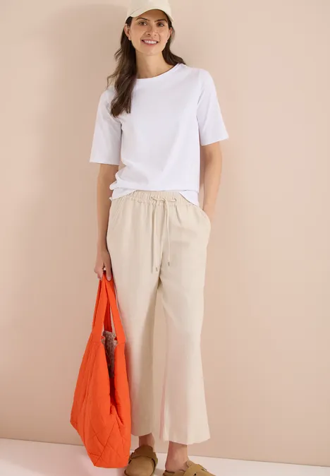 Basic Shirt met boothals White Vrouw in witte T-shirt en beige wijde broek met oranje tas, staande voor een beige achtergrond.