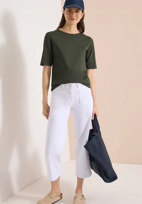 Basic Shirt met boothals khaki Vrouw in groen T-shirt met witte 7/8 wijdvallende broek, houdend een donkerblauwe jas in een minimalistische setting.
