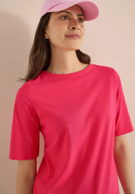 Basic Shirt met boothals geranium red Een persoon draagt een roze t-shirt en een bijpassende pet tegen een lichte achtergrond.