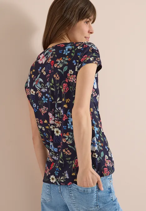 Eine Frau trägt ein kurzes, dunkelblaues Shirt mit buntem Blumenmuster und Jeans, seitlich stehend.