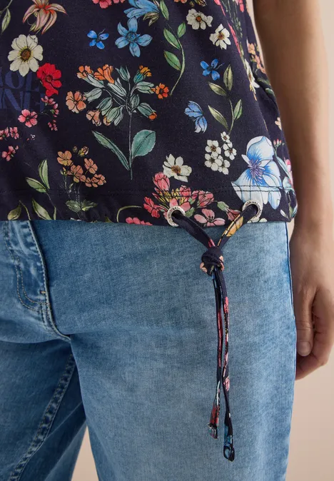 Schwarzes Top mit buntem Blumenmuster und Kordelzug am Saum, kombiniert mit blauen Jeans.