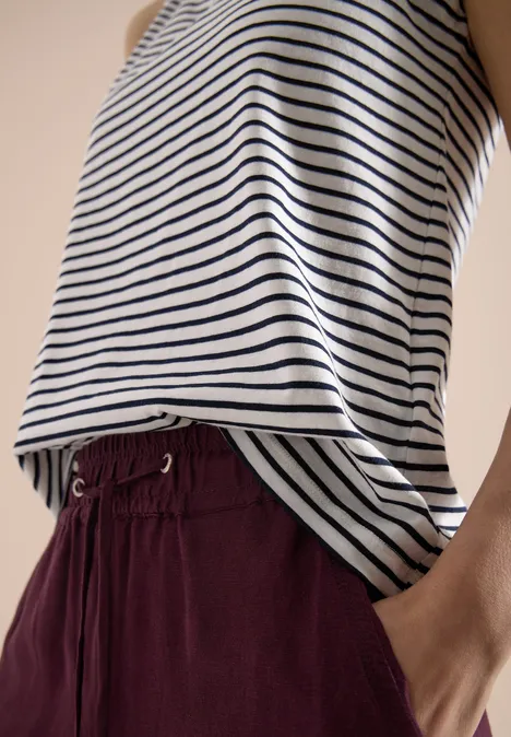 Bluzka w biało-granatowe paski, bordowe spodnie ze ściągaczem, ręka w kieszeni.