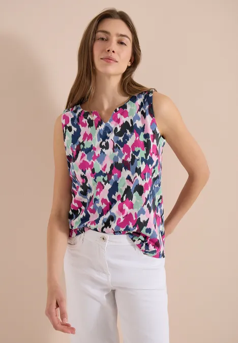 Femme en haut sans manches coloré à motif abstrait et pantalon blanc, fond neutre.