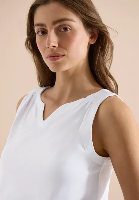 Vrouw in een witte mouwloze top met V-hals, opzij kijkend.