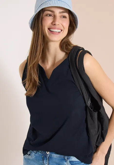 Frau in ärmellosem, dunkelblauem Oberteil trägt Rucksack und Hut, steht seitlich vor hellem Hintergrund.