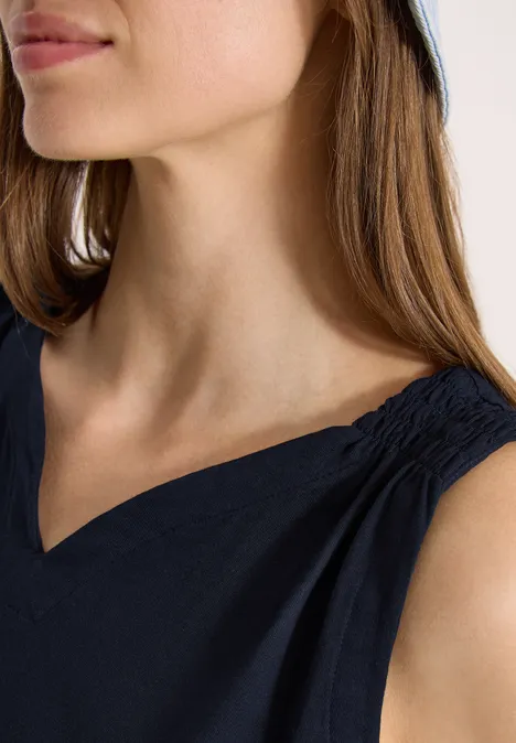 Ansicht einer Frau mit langem braunem Haar, hellem Hut, marineblauer V-Ausschnitt-Bluse mit gerafften Schultern.