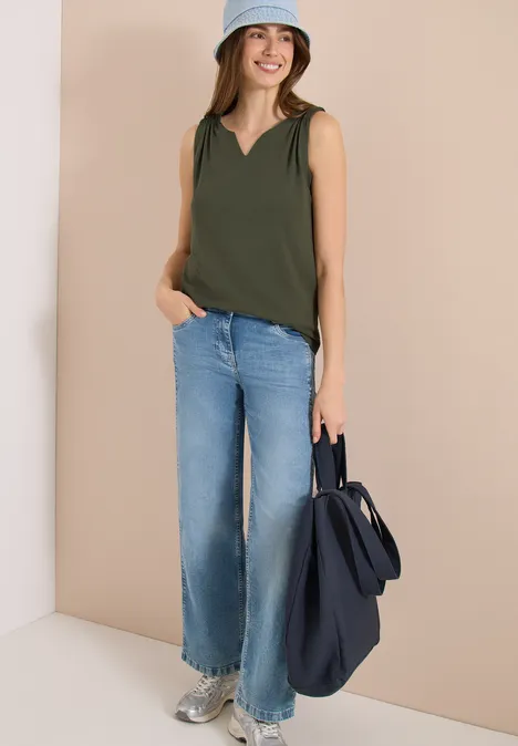 Een vrouw draagt een mouwloze groene top, lichte wijde jeans en houdt een donkerblauwe tas vast.