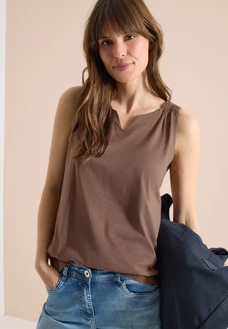 Vrouw draagt een bruin mouwloos topje en jeans, met een jas in de hand.