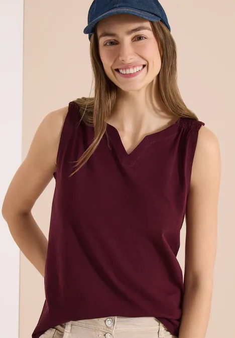 Vrouw met een bordeaux mouwloze top en een blauwe baseballpet voor een lichte achtergrond.