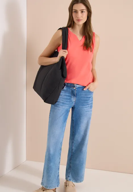 Vrouw met rood mouwloos topje, wijde blauwe jeans en grote zwarte tas voor een neutrale achtergrond.