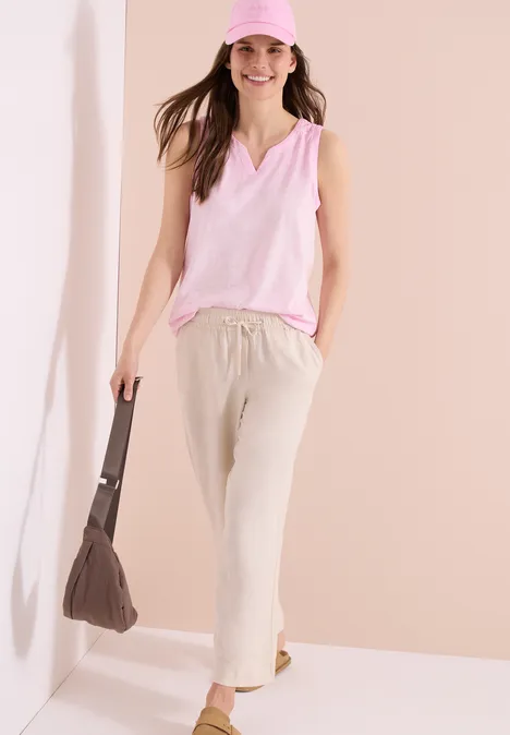 Frau in rosa ärmellosem Top und beigen Leinenhosen, Keilabsatz-Sandalen und brauner Tasche vor hellem Hintergrund.