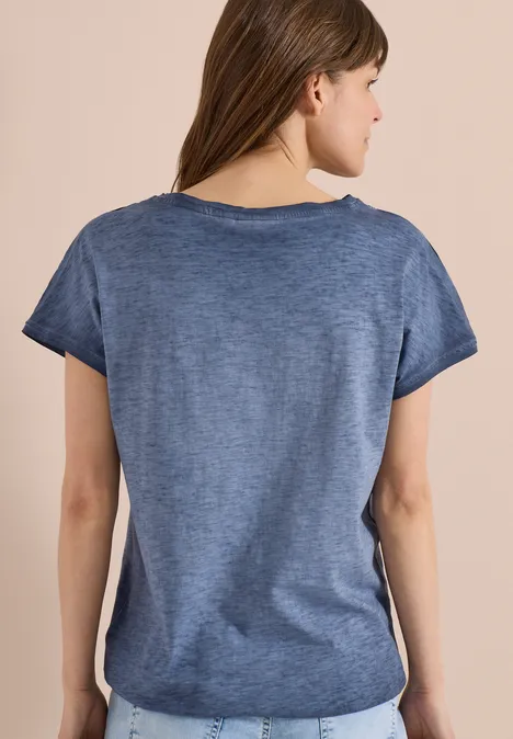 Rückansicht einer Frau mit kurzem blauen T-Shirt und abgerundetem Saum vor beigem Hintergrund.