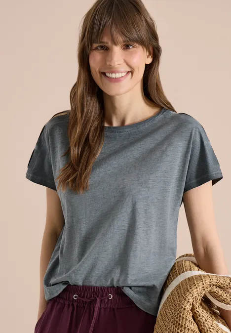 Vrouw in losse grijze t-shirt, deels ingestopt, houdt een beige jas in haar hand.
