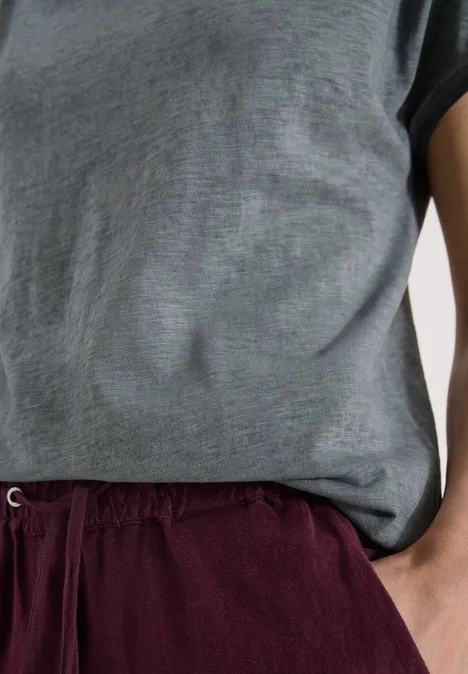 Bovenlichaam met grijs T-shirt en bordeauxrode short, persoon staat ontspannen met hand in zak.