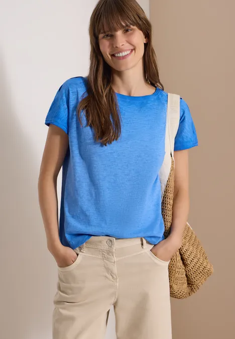 Frau mit blauem T-Shirt, beiger Hose und Strohtasche steht vor neutralem Hintergrund.