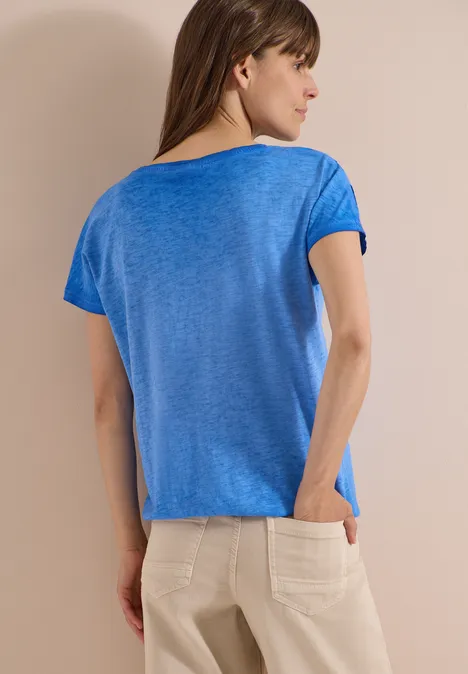 Eine Frau von hinten in blauem T-Shirt und beiger Hose, eine Hand in der Tasche.